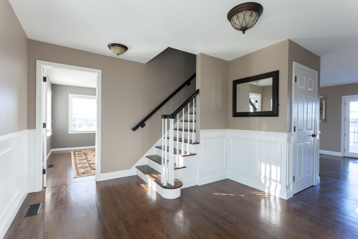 Bright hallway with hardwood floors, white wainscoting, and a wooden staircase with black handrail in a modern home.