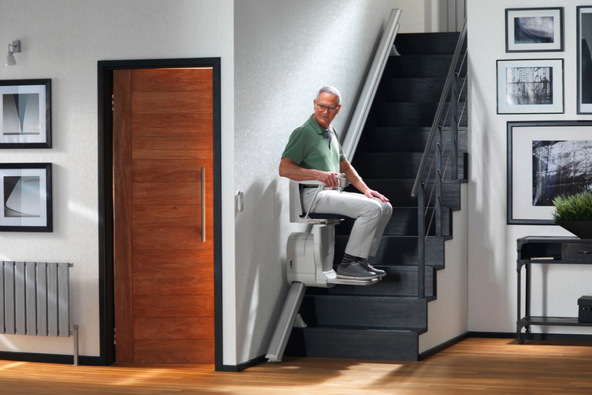 Older man in pyjamas seated on a straight stairlift on a dark modern staircase in a contemporary home.